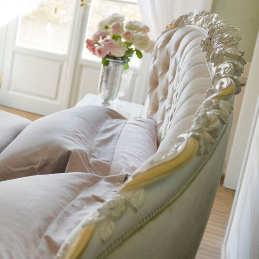 An elegant, vintage-style sofa featuring a tufted backrest and intricate floral carvings on a white wooden frame. It is beautifully adorned with soft pastel-colored cushions and positioned near a window. This setup exudes the charm of an Elegant Button Upholstered Winged Bed, complemented by a glass vase filled with pink and white flowers placed on a table in the background.