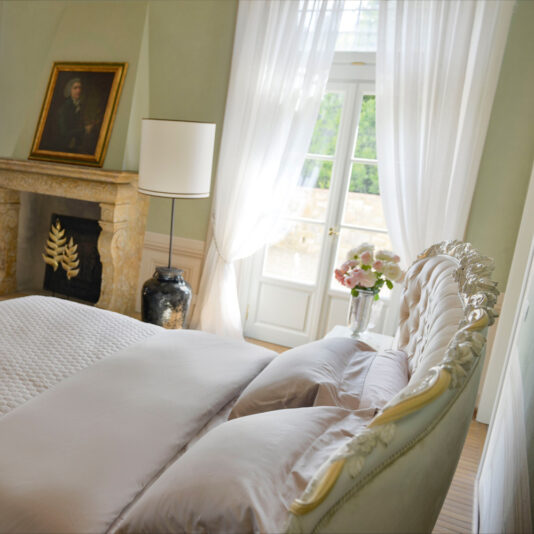 Luxurious bedroom with an Elegant Button Upholstered Winged Bed, soft white bedding, and a tufted headboard. A large window with sheer white curtains allows natural light to fill the room. A fireplace with an elegant mantel features a framed portrait. A vase of pink flowers sits by the window.