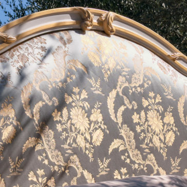 Elegant Classic Style Bed The Elegant Classic Style Bed boasts an ornate, gold-patterned headboard adorned with intricate floral designs set against a light background. Detailed carvings grace the top edge, and sunlight casts gentle shadows that enhance the depth and luxurious craftsmanship of this elegant piece.