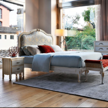 Elegant Classic Style Bed A luxurious bedroom with the Elegant Classic Style Bed, featuring an ornate headboard and a soft blue bedspread adorned with red accent pillows. A matching intricate nightstand, graced by a coral decoration, stands next to the bed. The room boasts large windows, wooden floors, and a scenic outdoor view.