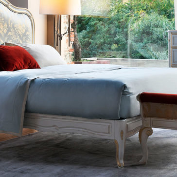 Elegant Classic Style Bed A serene bedroom with the Elegant Classic Style Bed dressed in light blue sheets and accented by a red pillow. The bed features an ornate, white and gold headboard. A cushioned bench sits at the foot of the bed. A floor lamp and large window with a view of greenery complete the room.