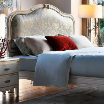 Elegant Classic Style Bed A luxurious bedroom showcasing the Elegant Classic Style Bed, highlighted by an ornate, upholstered headboard covered in sophisticated pale blue and gold damask fabric. The bed is dressed with light blue sheets and accented with a striking red throw pillow. A bedside table featuring a lamp and decorative branches completes the enchanting scene.