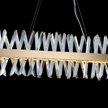 The Narrow Brass Crystal Chandelier features interconnected, zigzagging crystal panels and a long, horizontal gold bar at its center. The juxtaposition of its crystals and metal elements creates a striking modern aesthetic against a black background.