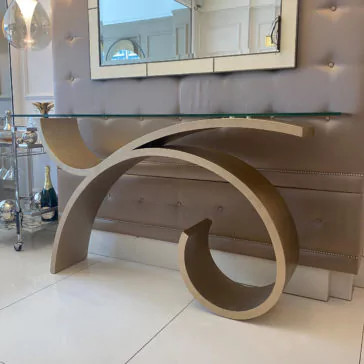 The Exclusive Champagne Leaf Console Table, with its modern design and artistic gold metal base featuring abstract curves and a glass top, adds elegance when positioned against a tufted, padded wall adorned with mirrored tiles in a sleek, contemporary interior. This sophisticated setting is further enhanced by the presence of a beverage cart on the left.