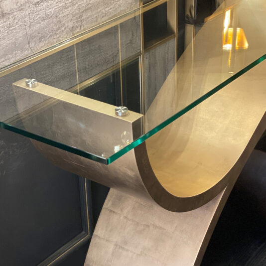 A modern glass table with a clear top supported by an artistic, curved metallic base, similar in style to the Exclusive Champagne Leaf Console Table. Positioned against a textured wall with reflective panels, it creates a contemporary and sleek aesthetic. A glowing lamp is partially visible in the background.
