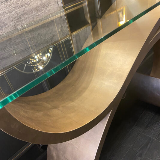The Exclusive Champagne Leaf Console Table features a sleek, modern design with a unique curved metallic base and a transparent glass top. Set against a textured wall with a geometric pattern, the chandelier's soft reflection shimmers on its pristine glass surface.