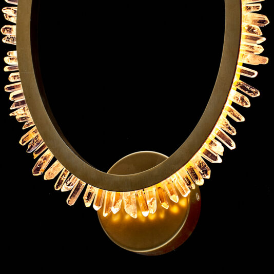 Exclusive Quartz Crystal Gold Wall Light The Exclusive Quartz Crystal Gold Wall Light is a unique circular wall-mounted lamp featuring jagged, quartz crystal-like light elements encircling the ring and emitting a warm glow. Its gold base blends seamlessly with the golden illumination from the crystals against a black background.