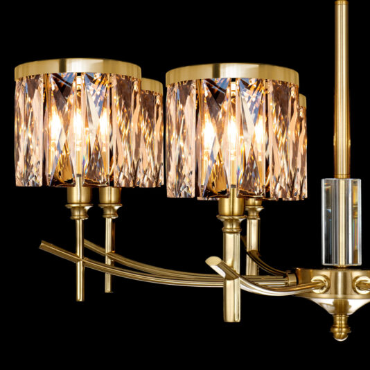 A close-up photograph of the High End 8 Arm Gold Chandelier showcases its two ornate, cylindrical lampshades made of faceted, golden-hued crystal. The fixture's elegant golden arms and accents cast a warm, radiant glow against a black background.
