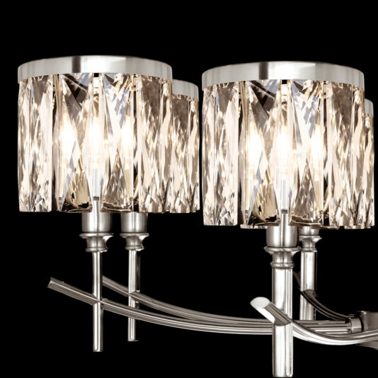 A close-up of a modern wall sconce featuring two cylindrical crystal lamp shades. The shades have intricate, faceted details that create a sparkling, light-reflecting effect. The sleek, metallic finish with curved, elegant arms evokes the sophistication of the High End 8 Arm Nickel Chandelier. The background is black.