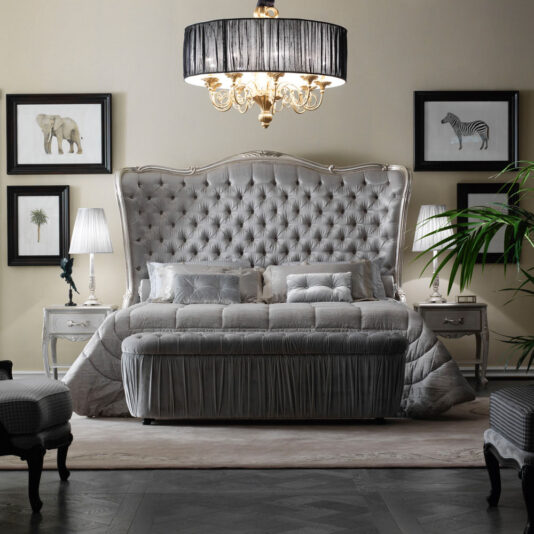 High End Button Upholstered Winged Bed A luxurious bedroom featuring the High End Button Upholstered Winged Bed with a plush, tufted headboard, expertly complemented by a neatly made bed and a matching bench at its foot. The room is adorned with a grand chandelier, elegant bedside tables with lamps, and wall art depicting an elephant, zebra, and plant.
