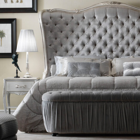 High End Button Upholstered Winged Bed A luxurious bedroom featuring a High End Button Upholstered Winged Bed adorned with an opulent, tufted headboard. The sophisticated bed is dressed in matching gray bedding and multiple pillows. To the left, a white nightstand holds a silver lamp and a decorative item. Framed artwork hangs on the wall in the background.