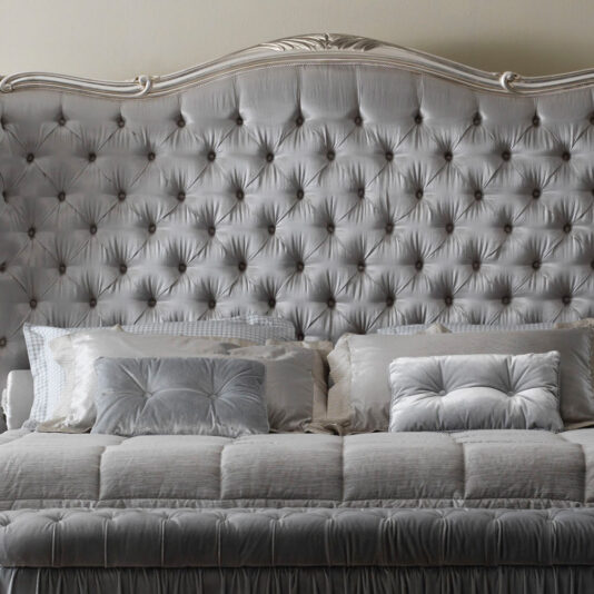 High End Button Upholstered Winged Bed The High End Button Upholstered Winged Bed features a sumptuous, tufted silver-grey headboard adorned with an elaborate, carved top edge. When combined with a coordinating bed set and an assortment of pillows in diverse shades of grey and silver, it forms a sophisticated and opulent bedroom arrangement.