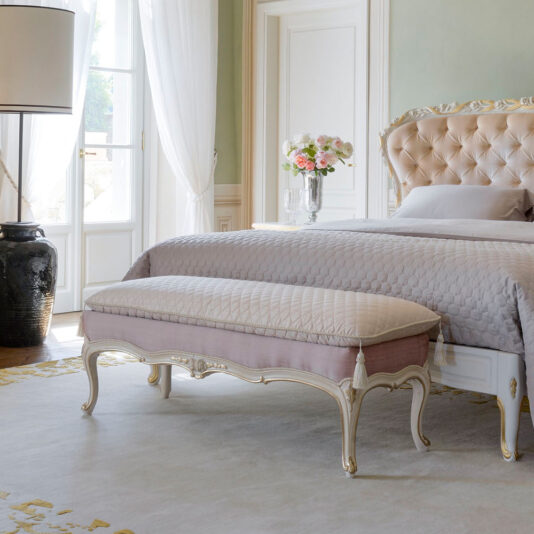 A beautifully decorated bedroom with a grand upholstered bed featuring a tufted headboard, draped in light purple and white bedding. In front of the bed is the Louis Style Classic Upholstered Bench. A large window with sheer curtains and a potted plant complete the elegant look.