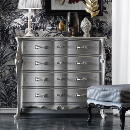 The Louis Style Tall Chest Of Drawers, with its silver metallic finish, curved drawers, and intricate handles, stands against a black and white jungle-themed wallpaper. A table lamp, decorative vases, and a golden leaf sculpture embellish the top. A checkered upholstered bench is partially visible.