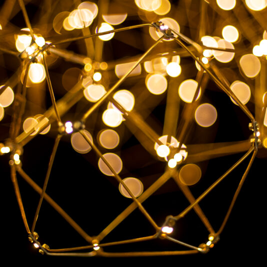 Luxury Set of 7 Prism Brass Pendant Lights Abstract image featuring a three-dimensional geometric wireframe structure with numerous out-of-focus warm yellow lights in the background, creating a bokeh effect. The wireframe structure resembles interconnected shapes, evocative of the Luxury Set of 7 Prism Brass Pendant Lights.