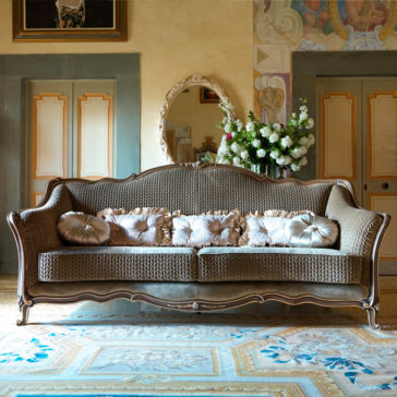 Luxury Venetian Style Sofa An elegant living room featuring the Luxury Venetian Style Sofa with ornate, patterned upholstery and several decorative pillows. Behind the sofa, there's a gilded mirror reflecting a lush floral arrangement. The room is adorned with art pieces and sophisticated architectural details.
