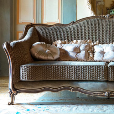 Luxury Venetian Style Sofa The Luxury Venetian Style Sofa boasts a carved wooden frame and exquisite velvet upholstery. It includes sumptuous, patterned cushions of various sizes and colors, complemented by a round, decorative pillow. The sofa is showcased in an elegant room adorned with ornate wall paneling and a patterned rug.
