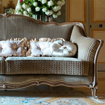 Luxury Venetian Style Sofa The Luxury Venetian Style Sofa, featuring an ornate wooden frame and plush, patterned upholstery, is adorned with several decorative silk pillows. Behind the sofa, a vase boasting a large bouquet of white flowers complements the elegant, paneled walls.