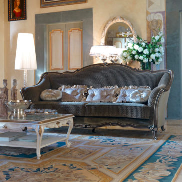 Luxury Venetian Style Sofa Elegant living room featuring the Luxury Venetian Style Sofa, adorned with plush pillows and a throw. The space includes a white ornate coffee table and tall floor lamps. A bouquet of white flowers is placed under an ornate mirror on a decorative wall, with a portrait hanging to the side.