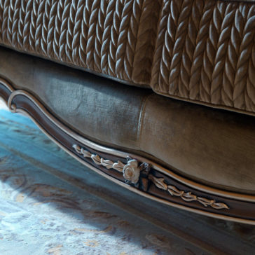 Luxury Venetian Style Sofa A close-up view of the intricately carved base of a bed with a detailed floral design reveals the exquisite craftsmanship. The fabric of the bedspread above showcases a woven pattern, while the floor is adorned with a patterned rug in muted tones. Adjacent to this setup is the Luxury Venetian Style Sofa, enhancing the overall elegant and luxurious atmosphere.