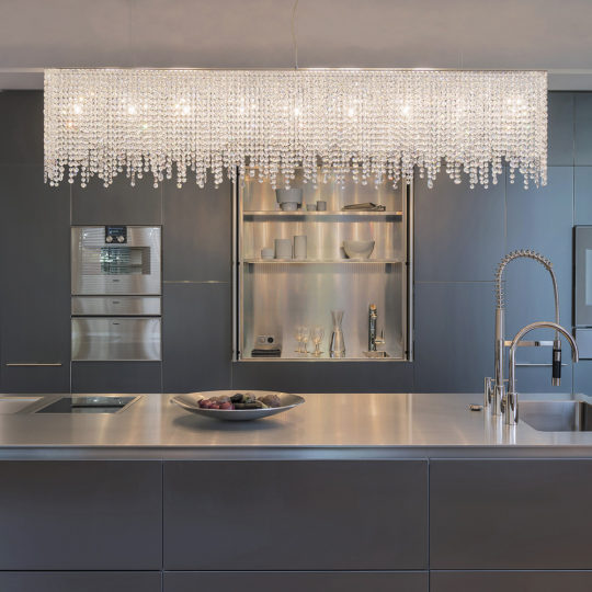 Modern kitchen with sleek dark cabinets and stainless steel appliances. A Deluxe Modern Narrow Crystal Chandelier hangs above a metallic kitchen island with an integrated stovetop and sink. A bowl with fruit is placed on the countertop.