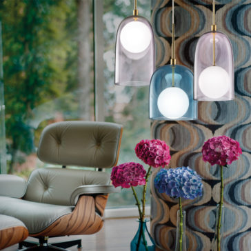 Contemporary Long Glass Dome Pendant Light A cozy living room features a modern armchair and three stylish Contemporary Long Glass Dome Pendant Lights in various vibrant shades. A tall vase filled with pink and purple hydrangeas is positioned on the floor, set against large windows that reveal a lush green forest.
