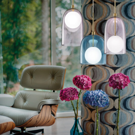 Contemporary Long Glass Dome Pendant Light A cozy living room features a modern armchair and three stylish Contemporary Long Glass Dome Pendant Lights in various vibrant shades. A tall vase filled with pink and purple hydrangeas is positioned on the floor, set against large windows that reveal a lush green forest.