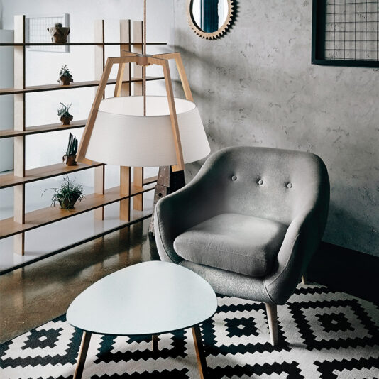 Modern Oak Frame Pendant Light A cozy, modern living space featuring a tufted gray armchair, a triangular white coffee table, and a black-and-white patterned rug. The Modern Oak Frame Pendant Light hangs uniquely above, with a minimalist bookshelf adorned with potted plants in the background.