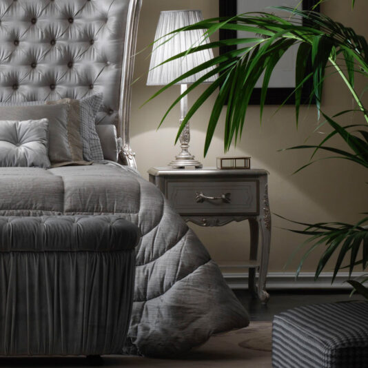 A cozy, elegant bedroom featuring a plush tufted headboard, a matching grey bedspread, and the Ornate Bedside Cabinet With Drawer holding a classic lamp. A potted palm adds a touch of greenery, while framed artwork adorns the walls, creating a serene and sophisticated ambiance.