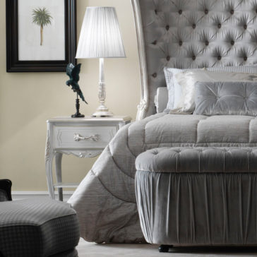 A luxurious bedroom features a tufted, silver headboard bed with grey bedding. The Ornate Bedside Cabinet With Drawer, holding a lamp and parrot statue, stands next to the bed. Above the cabinet hangs a framed palm tree artwork. A grey checkered ottoman is at the end of the bed.