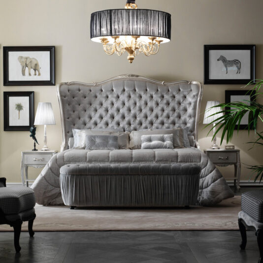 Elegant bedroom featuring a large, tufted headboard bed with plush bedding. Two Ornate Bedside Cabinets With Drawers and decorative lamps flank the bed. A chandelier with a black shade hangs overhead. Framed animal artwork adorns the wall, and a plant adds a touch of greenery.