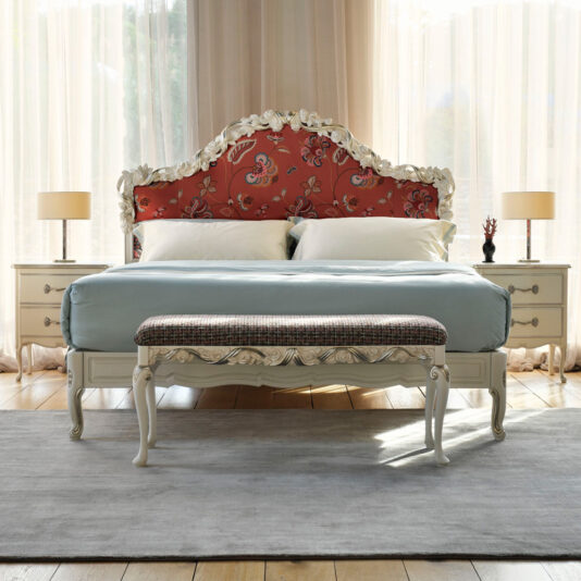 A bedroom featuring the Ornate Classic Style Upholstered Bed, which boasts a large, red floral-patterned headboard. The bed is adorned with light blue bedding and complemented by a cushioned bench at its foot. On either side of the bed are two white nightstands, each topped with a lamp. Light curtains let natural light flood into the classic-style room.