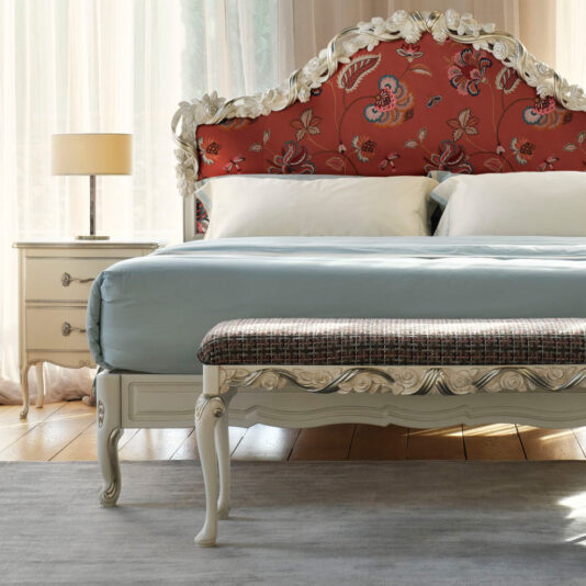 Elegant bedroom featuring the Ornate Classic Style Upholstered Bed, showcasing a plush design with a red, floral-patterned headboard and light blue bedding. At the foot of the bed sits a patterned bench. The room includes a white nightstand with a lamp, and natural light fills the space through sheer curtains, perfectly embodying an ornate classic style.