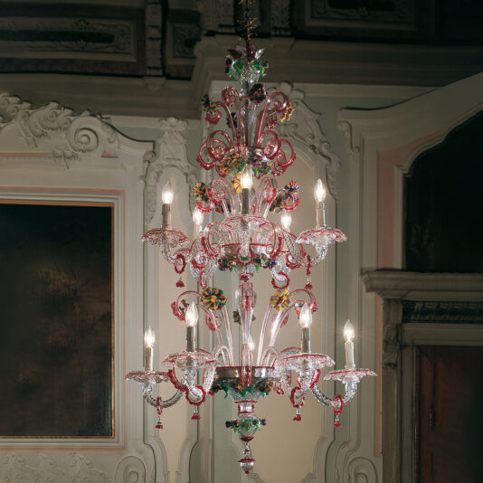 The Ornate Glass Chandelier With Red Floral Detailing, featuring pink accents and multiple glowing light bulbs, hangs from a ceiling in an elaborate, classic interior with detailed wall moldings. This chandelier showcases a mix of clear and colored glass elements.