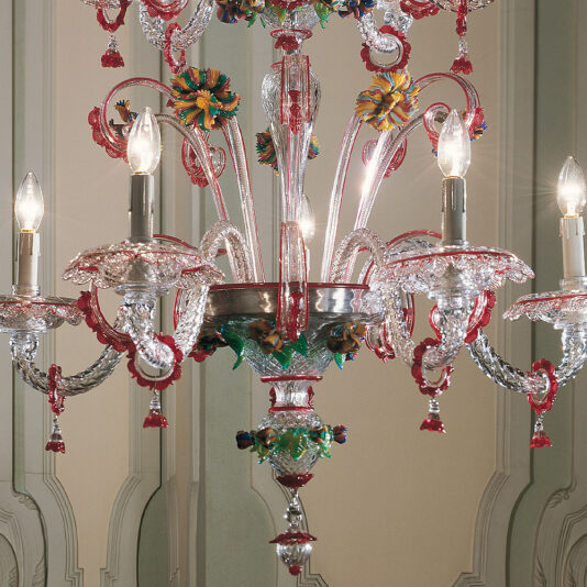 Ornate Glass Chandelier With Red Floral Detailing A close-up of the Ornate Glass Chandelier With Red Floral Detailing, showcasing intricately designed and colorful elements. The chandelier features multiple arms adorned with decorative details and lit candle-like bulbs, all set against a background of elegant wall paneling in a light, neutral color.