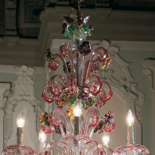 Ornate Glass Chandelier With Red Floral Detailing A close-up of the Ornate Glass Chandelier With Red Floral Detailing, showcasing its intricate glasswork and vibrant decorations. The light fixture features multiple candle-like bulbs encircled by swirling glass designs and is set against an elegantly adorned ceiling.