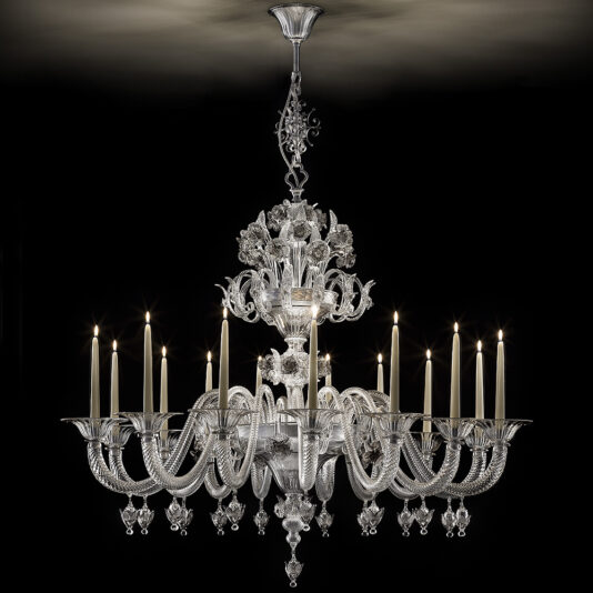 The Ornate Floral Glass Chandelier With Candle Details, suspended from the ceiling, showcases intricate floral designs. It boasts twelve candle-like lights arranged in a circular pattern, each encased in a detailed holder, casting a warm glow that illuminates the room against a dark background.