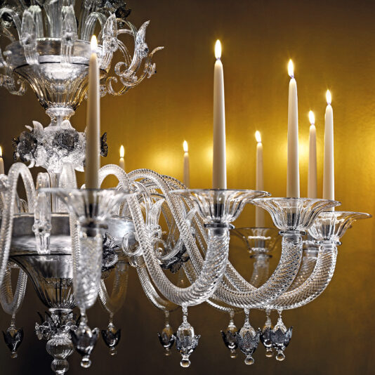 Ornate Floral Glass Chandelier With Candle Details A close-up of the Ornate Floral Glass Chandelier With Candle Details reveals its intricate design, featuring curving arms and decorative elements. Highlighted against a warm, gold-toned background, it casts an elegant and luxurious ambiance with flickering candlelight.