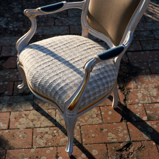 The Ornate Venetian Style Occasional Armchair, featuring a vintage wooden frame painted white with gold accents, dark blue padded armrests, and a cushioned seat covered in light patterned fabric, graces a weathered brick patio adorned with patches of moss.