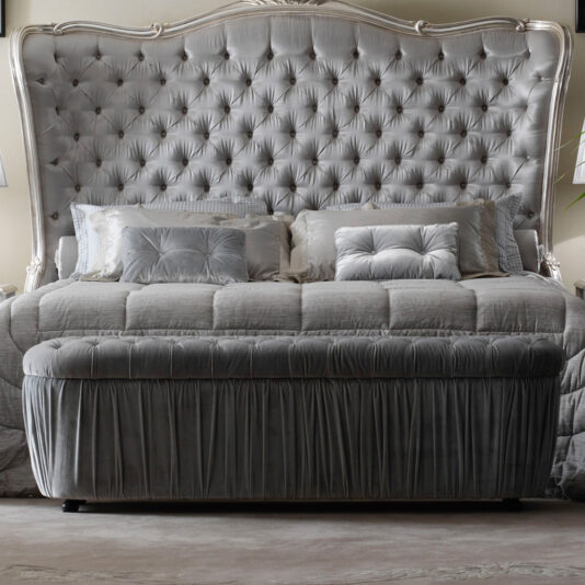 A luxurious bedroom features a large, plush bed with a tufted headboard. The bed is adorned with silver-gray bedding and multiple matching pillows. At the foot of the bed, there is an Ottoman Style Button Upholstered Storage Bench with a gathered skirt. The room is softly lit.