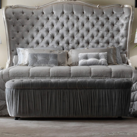 A luxurious bedroom features a large, plush bed with a tufted headboard. The bed is adorned with silver-gray bedding and multiple matching pillows. At the foot of the bed, there is an Ottoman Style Button Upholstered Storage Bench with a gathered skirt. The room is softly lit.