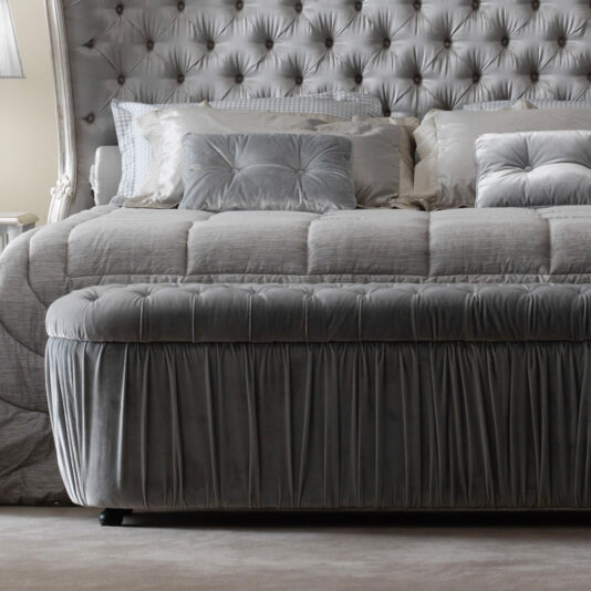 A luxurious, tufted headboard stands behind a well-made bed adorned with plush, grey pillows and a matching comforter. At the foot of the bed is an Ottoman Style Button Upholstered Storage Bench in the same grey fabric, all set in an elegantly designed bedroom.