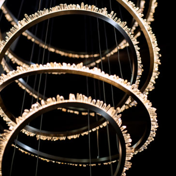 A close-up of the Quartz Crystal 7 Ring Nickel Chandelier showcases its modern design with multiple circular rings adorned with illuminated quartz crystals. The rings are suspended in a vertical sequence, creating a layered effect. The soft lighting enhances the intricate design against a dark background.