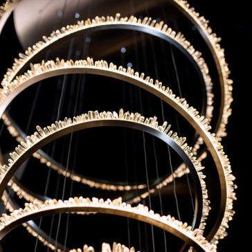 A close-up view of the Quartz Crystal 7 Ring Nickel Chandelier, featuring seven vertically arranged circular rings in a sleek nickel finish. Each ring is embellished with numerous small crystal-like elements that emit a warm, glowing light, crafting an elegant and intricate design against a dark background.