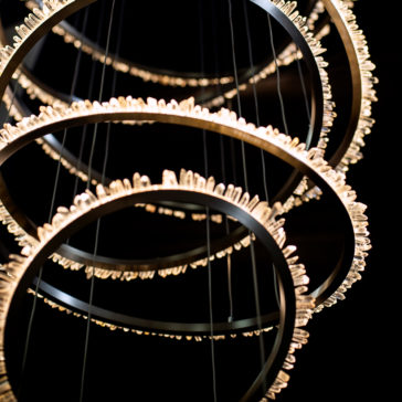 The Quartz Crystal 7 Ring Nickel Chandelier is a contemporary lighting fixture featuring multiple circular rings arranged in a staggered formation. Each ring is adorned with small lights along its circumference, casting a radiant and elegant pattern against a dark backdrop.