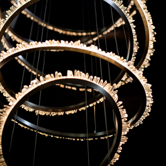 Quartz Crystal 7 Ring Nickel Chandelier The Quartz Crystal 7 Ring Nickel Chandelier is a contemporary lighting fixture featuring multiple circular rings arranged in a staggered formation. Each ring is adorned with small lights along its circumference, casting a radiant and elegant pattern against a dark backdrop.
