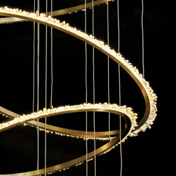 Quartz Halo 7 Ring Gold Chandelier A close-up of the Quartz Halo 7 Ring Gold Chandelier showcases its interlaced circular bands embellished with tiny, radiant lights. The chandelier hangs from several vertical wires, standing out against a dark backdrop and highlighting its detailed and graceful design.
