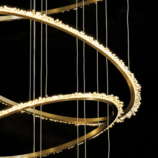 Quartz Halo 7 Ring Gold Chandelier A close-up of the Quartz Halo 7 Ring Gold Chandelier showcases its interlaced circular bands embellished with tiny, radiant lights. The chandelier hangs from several vertical wires, standing out against a dark backdrop and highlighting its detailed and graceful design.