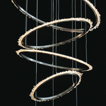 The Quartz Crystal 7 Ring Nickel Chandelier is an artistic lighting fixture featuring multiple circular, metal rings suspended in a spiral arrangement. Adorned with small, glowing lights along its edge, it creates a cascading effect against a dark background.
