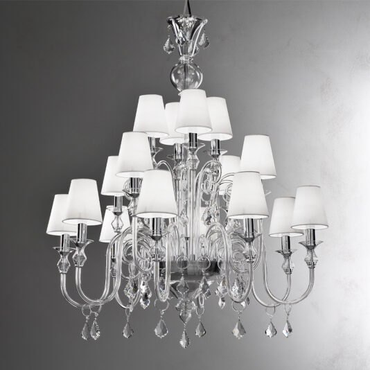 The Traditional Chandelier With Crystal Drops is a luxurious fixture featuring multiple tiers adorned with white lampshades and hanging crystal drops. Its intricate design boasts curved arms and an elegant central structure, all beautifully illuminated against a plain gray background.